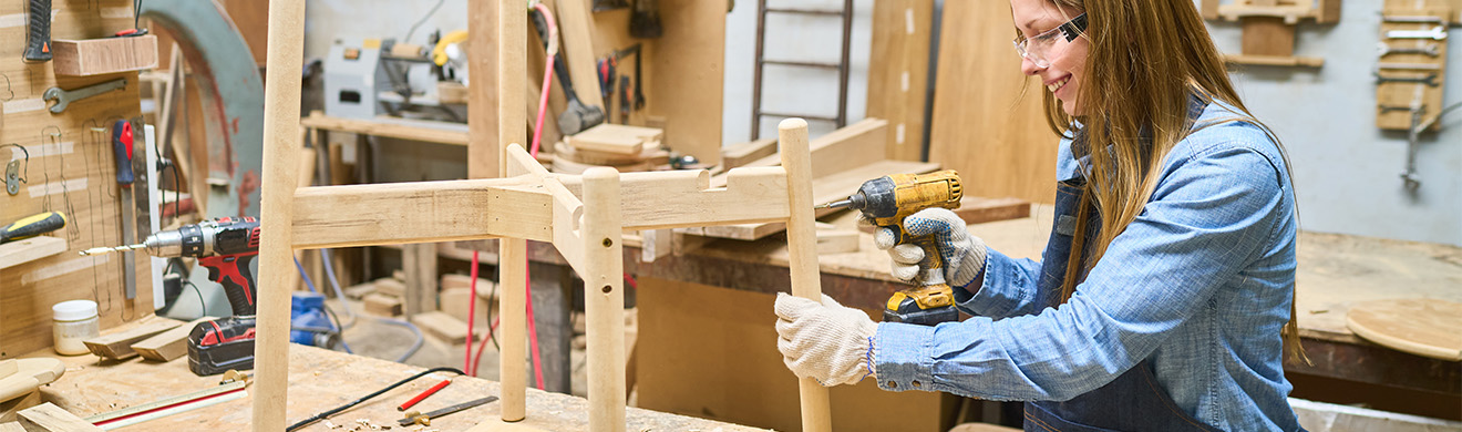 Junge Schreinerin montiert Holzmöbel in einer Werkstatt und arbeitet konzentriert mit Werkzeugen am Tisch
