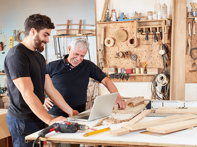 Senior und Lehrling besprechen Schreinerarbeiten in Euskirchen am Laptop, planen Holzprojekte und handwerkliche Aufgaben gemeinsam.