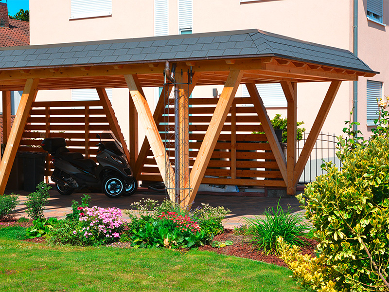 Moderner Carport aus lasiertem Holz mit Kupferdachrinne und offenem Einfahrbereich vor einem Wohnhaus in alpiner Architektur